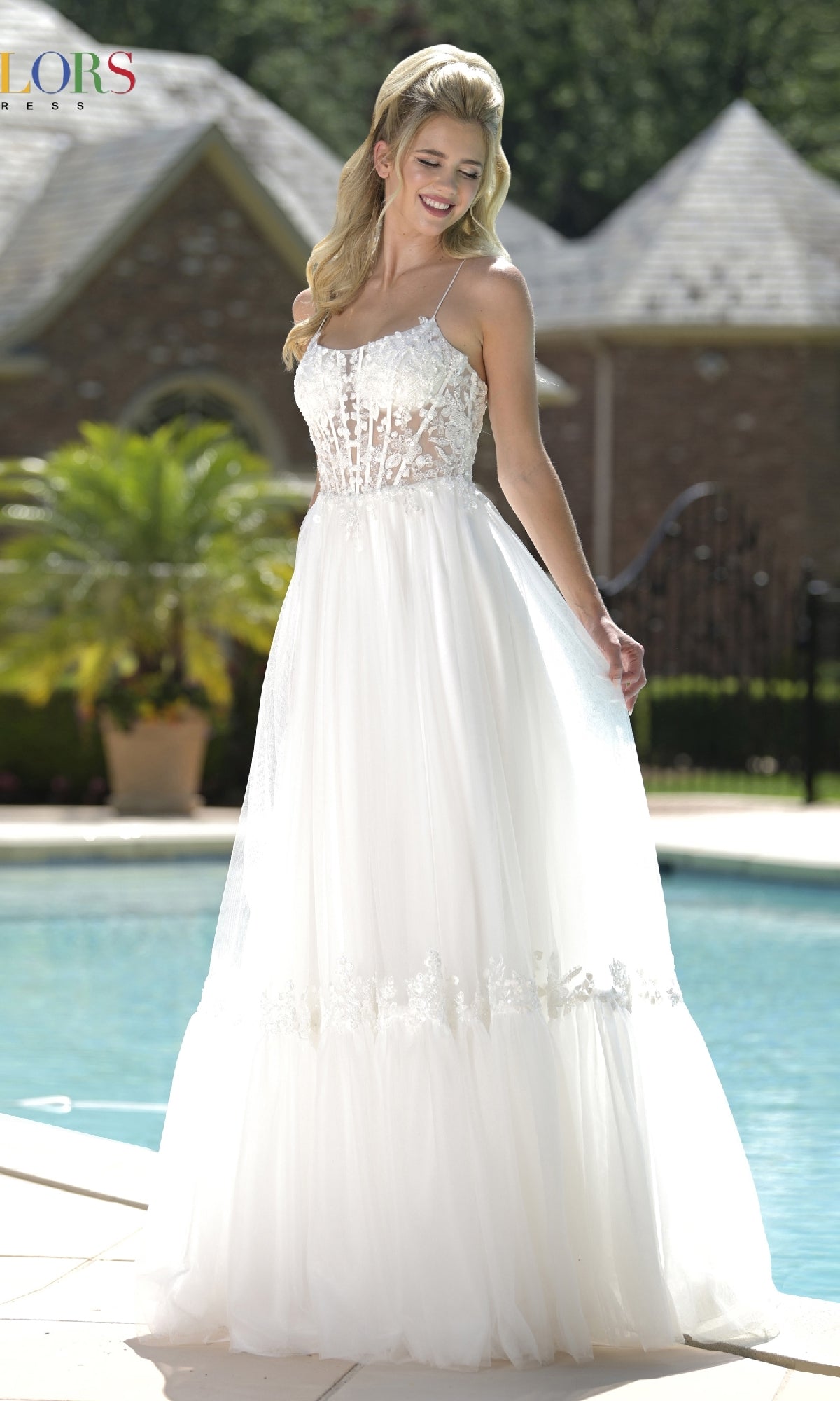 A woman in the Colors Dress 3799, a white sleeveless long prom dress with an embroidered sheer bodice, stands smiling by a pool with a large house and lush greenery behind her.